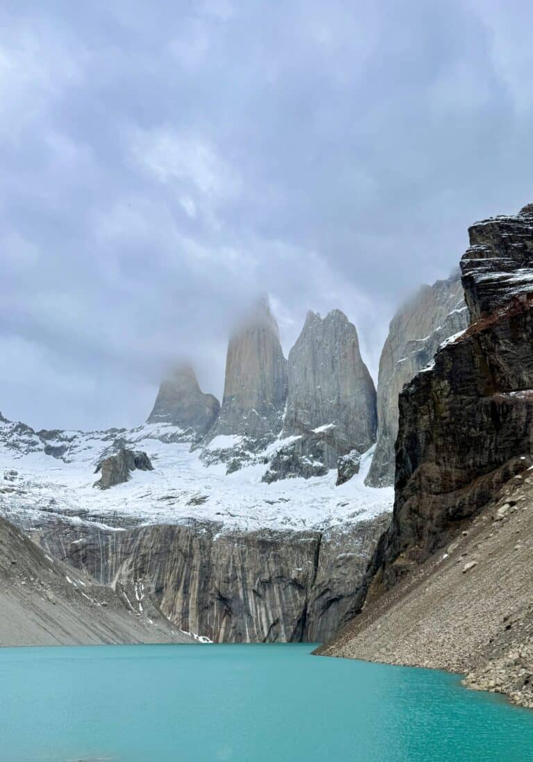 The three granite towers of Torres del Paine rising above snow and glacial rock under stormy clouds, reflected in the turquoise waters of Laguna de los Tres, Chilean Patagonia