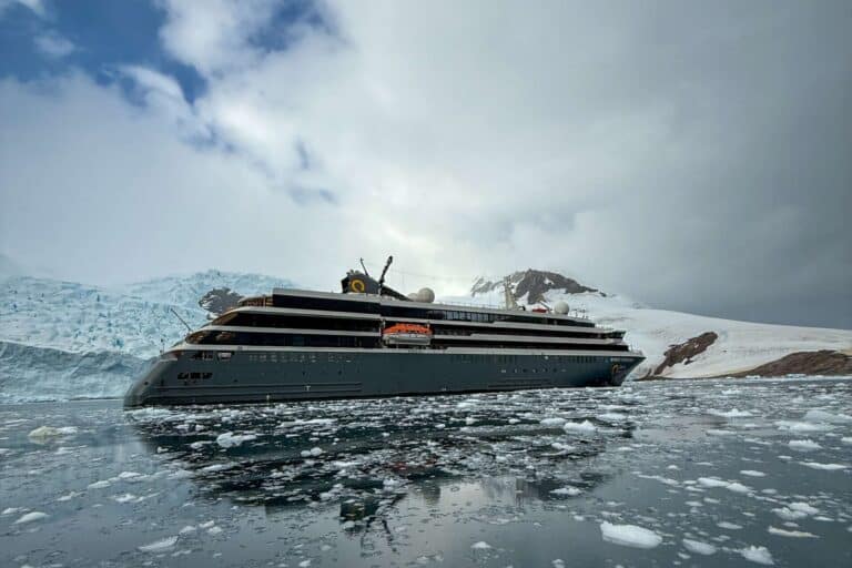 Expedition ship surrounded by brash sea ice in Antarctic waters, glacier face and snow-covered peaks visible behind