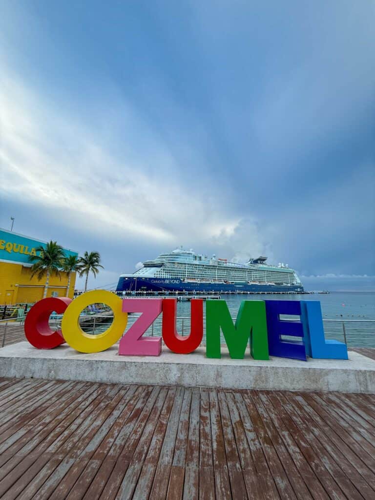 Colorful large-letter COZUMEL sign on a wooden pier with the Celebrity Beyond cruise ship docked in the harbor behind it, Cozumel, Mexico