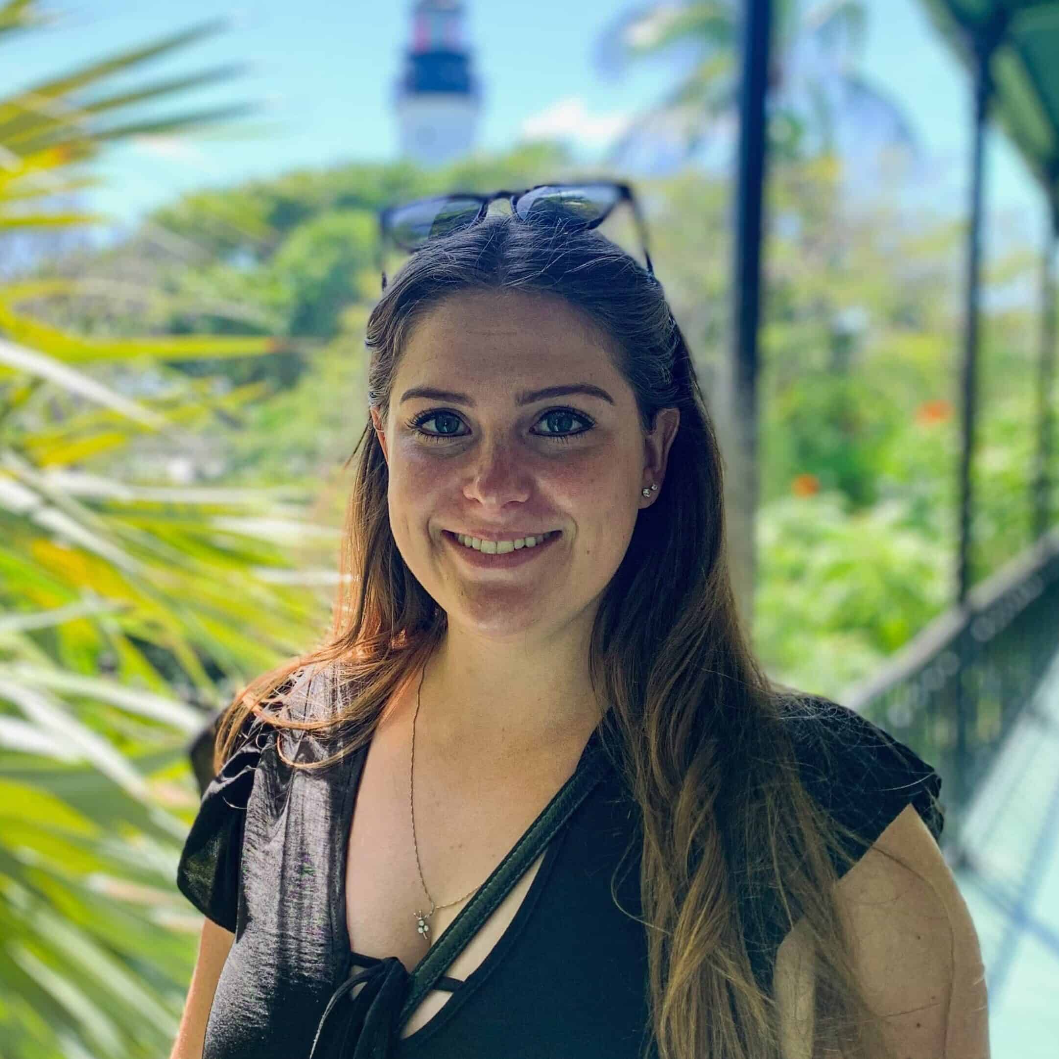 Kaitlin smiling on a tropical veranda with lush greenery and a historic lighthouse behind, Key West, Florida