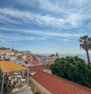 Rooftop terrace cafe overlooking Alfama district with terracotta rooftops, a palm tree, and the Tagus River in Lisbon, Portugal