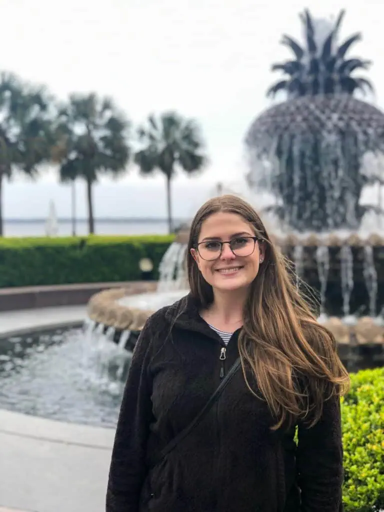 Kaitlin smiling in front of the Pineapple Fountain at Waterfront Park in Charleston, South Carolina, with palm trees and the harbor visible behind her