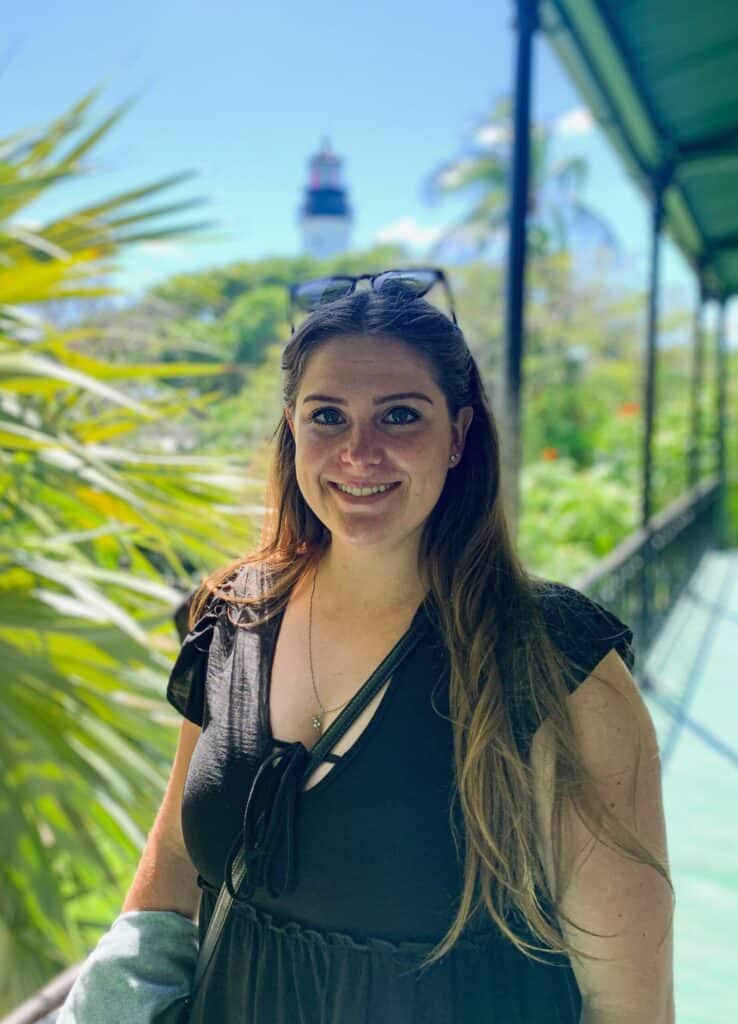 Kaitlin smiling on a tropical veranda with lush greenery and a historic lighthouse behind, Key West, Florida