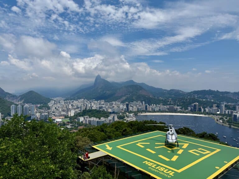 Aerial view from Sugarloaf Mountain helipad over Rio de Janeiro, with Christ the Redeemer visible on Corcovado mountain, Botafogo beach curving below, and the city skyline behind