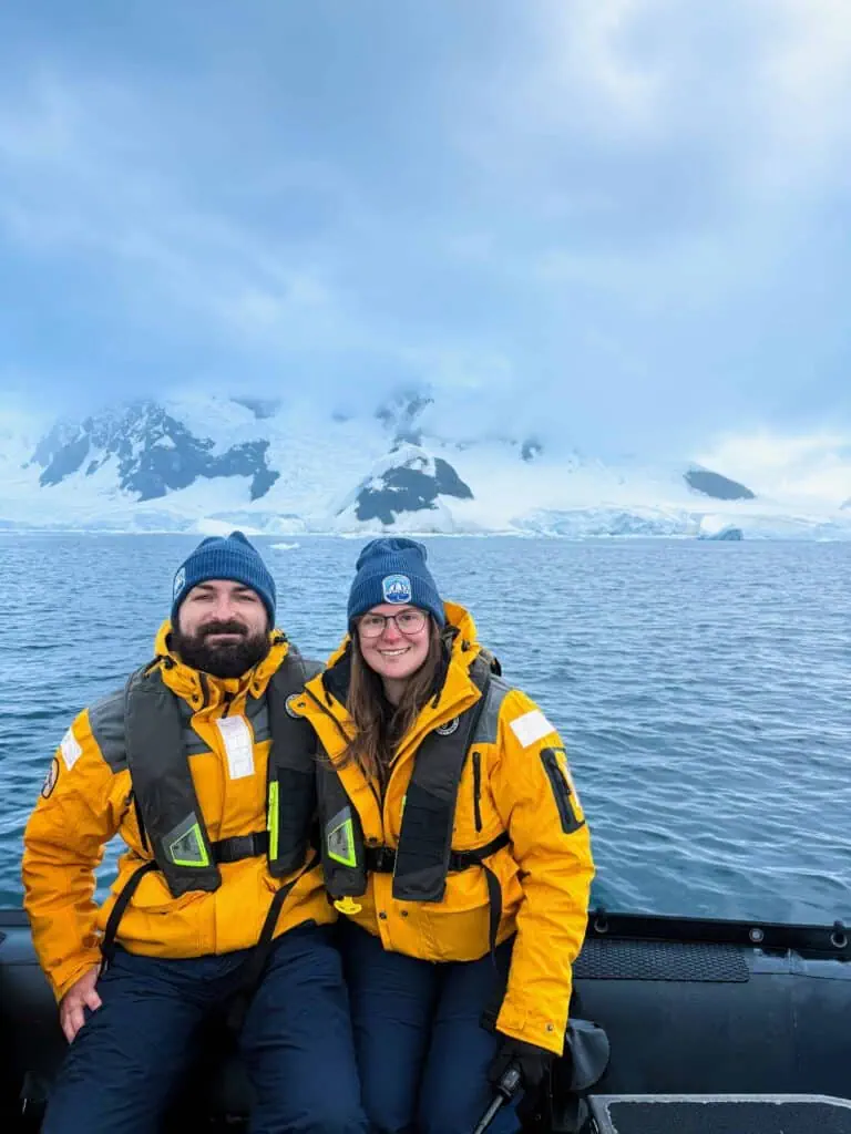 Kaitlin and her partner in bright yellow expedition jackets seated in a zodiac inflatable boat on calm Antarctic waters, with glaciers and snow-covered mountain peaks behind them