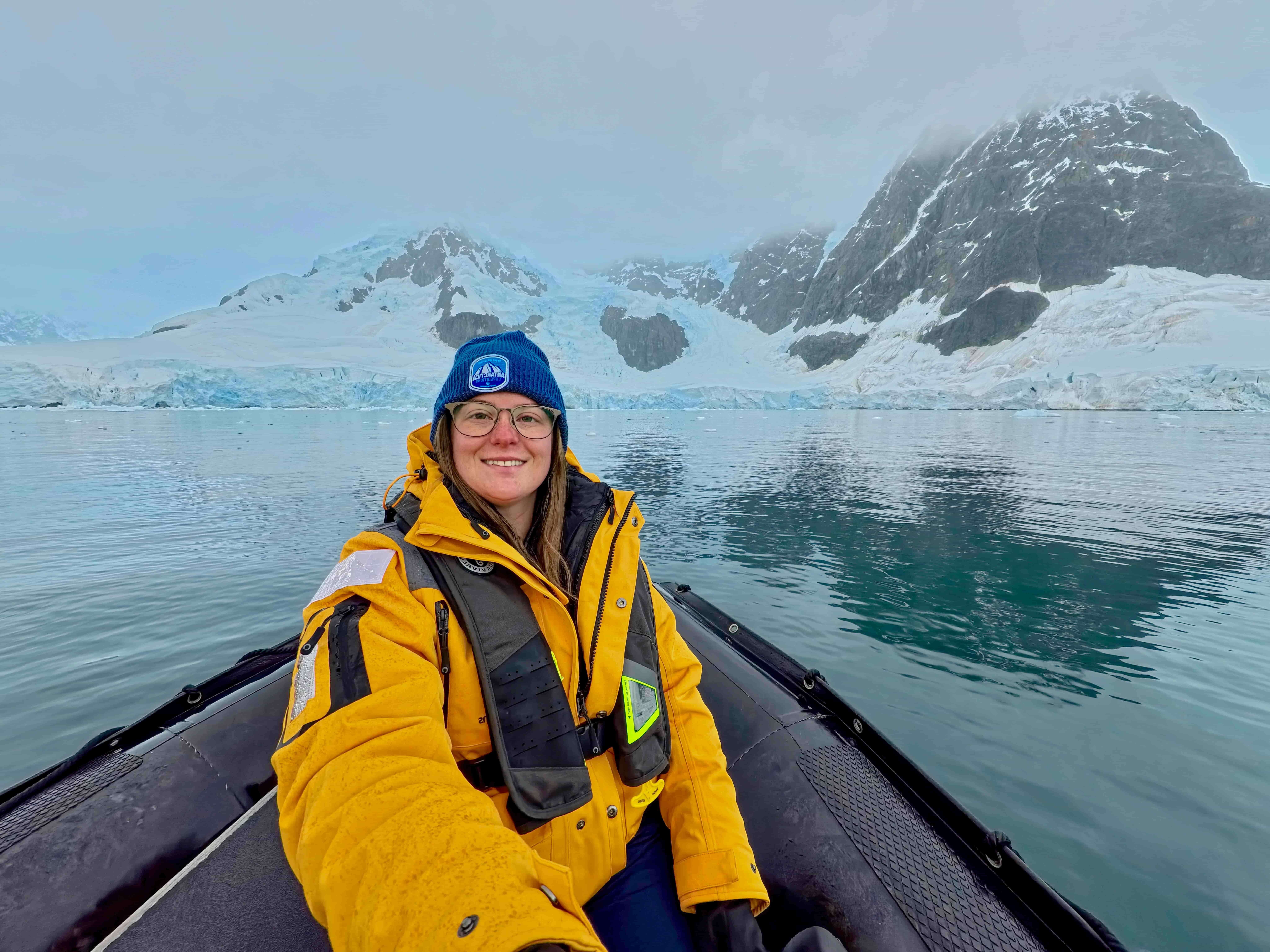 A traveler in a yellow expedition parka and life vest smiling on a zodiac in Antarctica with glaciers and snow-covered peaks in the background.