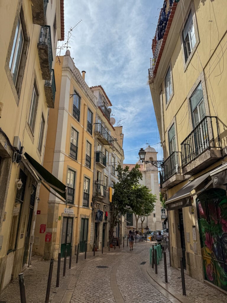 Narrow Alfama street in Lisbon leading toward a church tower, lined with restaurants including a fado house.