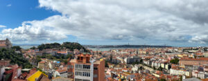 Panoramic view over Alfama's tiled rooftops toward the Tagus River, São Jorge Castle on the hill and the 25 de Abril Bridge in the distance.
