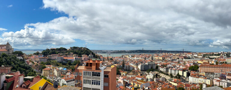 Panoramic view over Alfama's tiled rooftops toward the Tagus River, São Jorge Castle on the hill and the 25 de Abril Bridge in the distance.